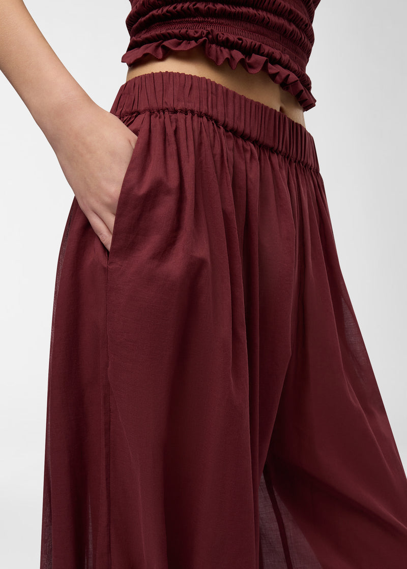 Detail view, Noor Maxi Pants: model places hand in side slit pocket, showing elastic low-rise waistband and semi-sheer, wide balloon legs in rouge noir.