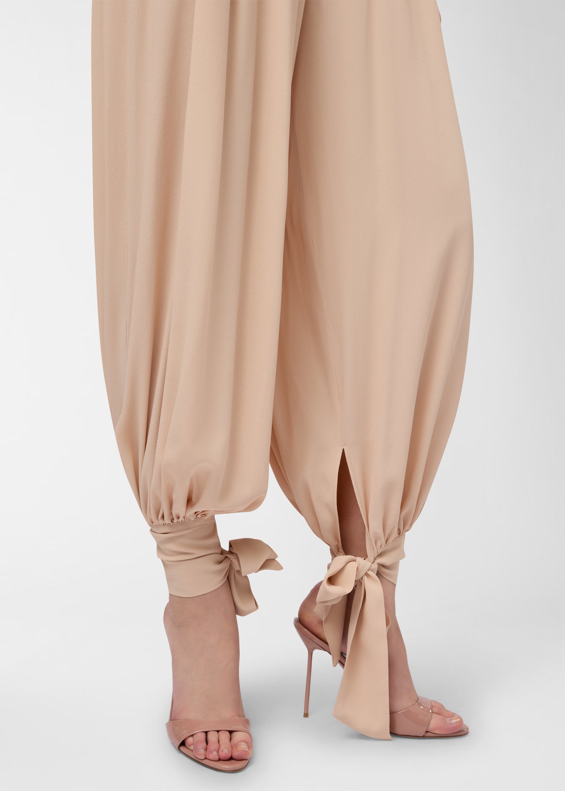 Detail view — Noor Maxi Pants: peach crepe-de-chine pants flowing in a wide balloon silhouette, tapering and tied in bows at the ankles over stiletto sandals.