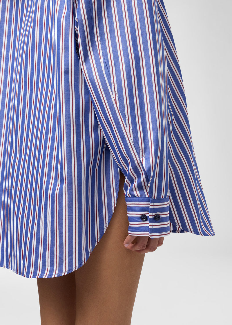 Detail view — New Georgiana Shirt: oversized blue and rouge noir maxi-striped cotton poplin, showing buttoned cuff, curved hem and side slit as it falls beside model’s thigh.