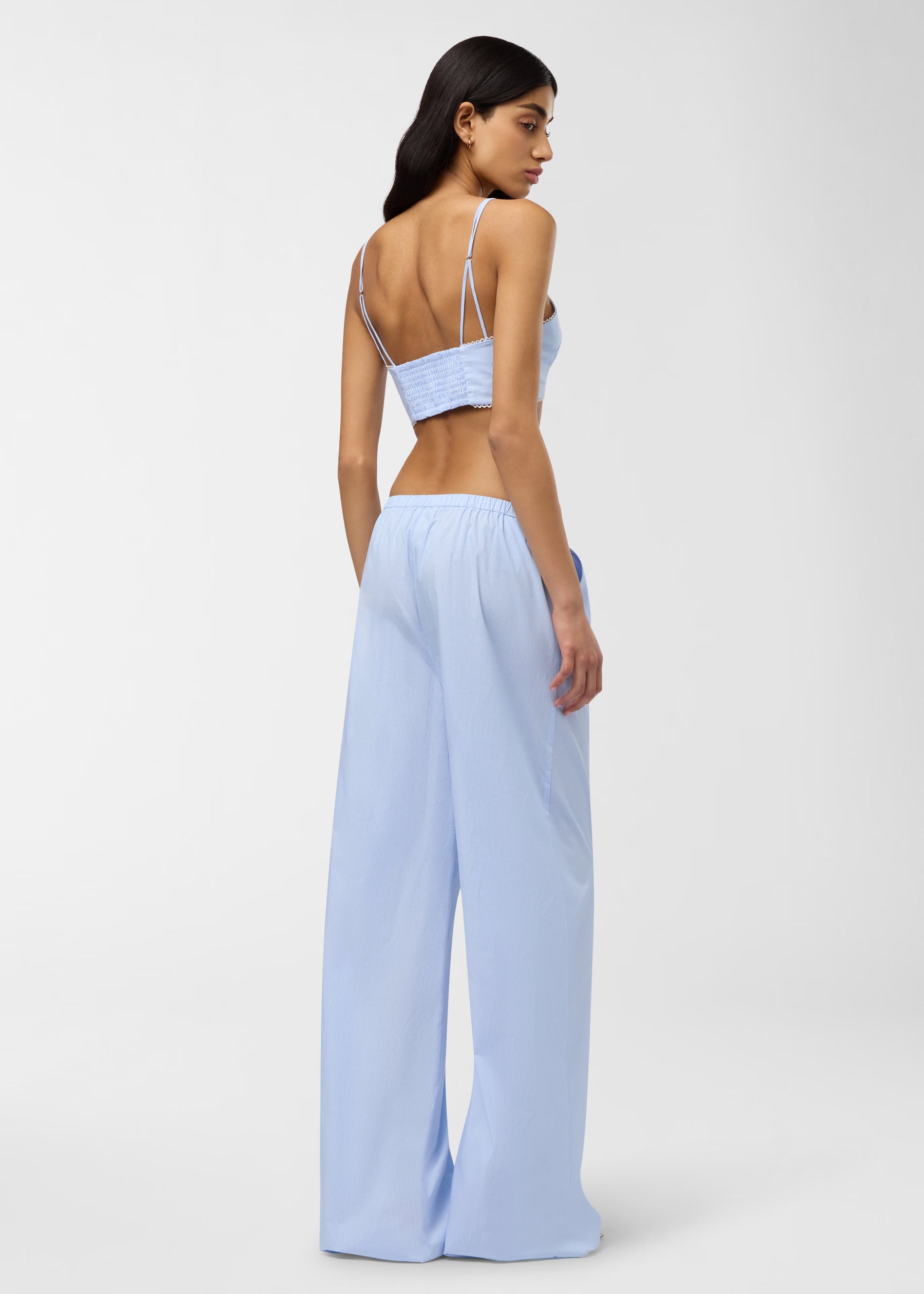 Rear view — Carrie Top: model wears a light blue and white micro-striped super-cropped bralette top with double spaghetti straps, ruched back and micro-lace hem, studio.