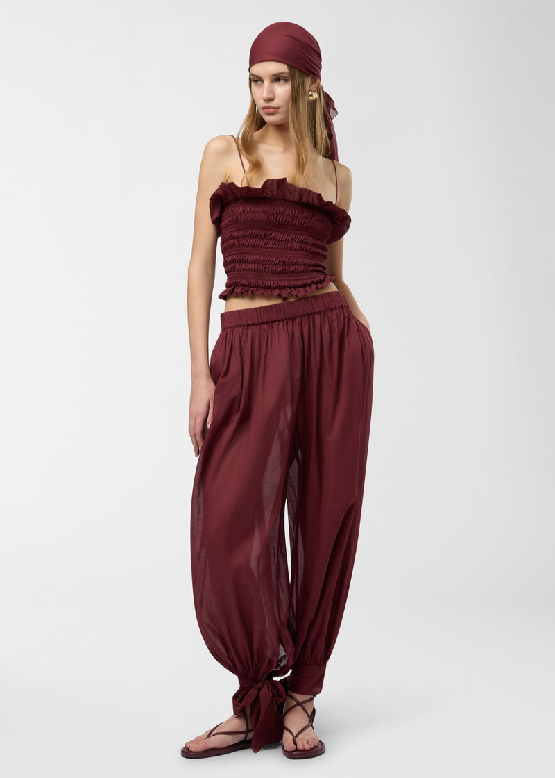 Frontal view — Model wearing Suri Top: rouge noir smocked tube top with undulated ruffle and spaghetti straps, paired with matching sheer tie-hem trousers and headscarf on white background.