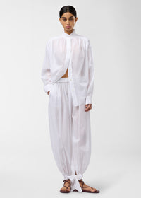 Frontal view — Rania Shirt: model wearing oversized white cotton muslin shirt, standing against a plain light background, mandarin collar, button placket, pleated shoulders and relaxed fit.