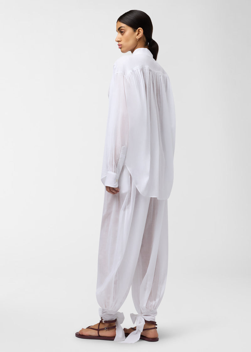 Rear view: Rania Shirt drapes on a model standing turned, showcasing semi-sheer white cotton muslin, mandarin collar, gathered pleats at shoulder and back yoke, and relaxed oversized fit.