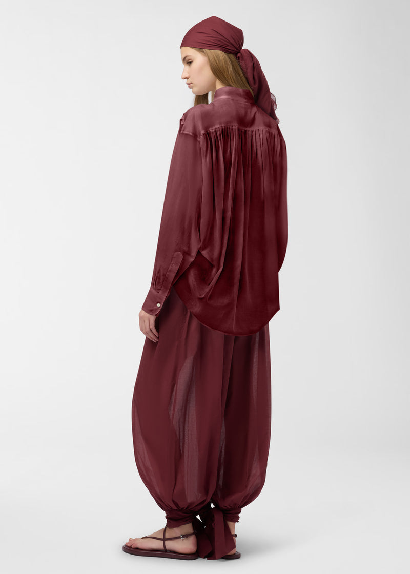 Rear view — Rania Shirt: model wears rouge noir oversized cotton-muslin shirt draping loosely, highlighting pleats along the shoulder and back yoke, mandarin collar and long cuffs in studio.