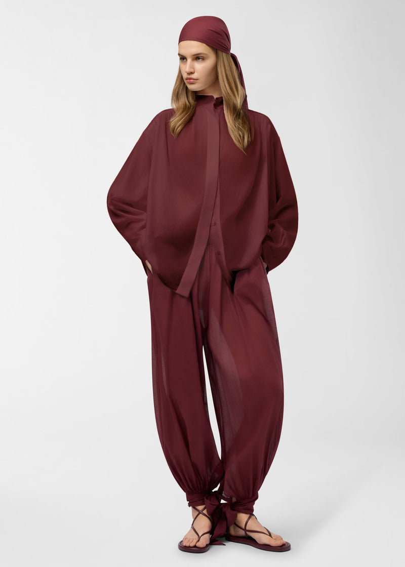 Frontal view — Model wears Rania Shirt in deep rouge noir, oversized semi-sheer cotton muslin with mandarin collar, concealed button placket, pleated shoulder and cuff details; hands in pockets.