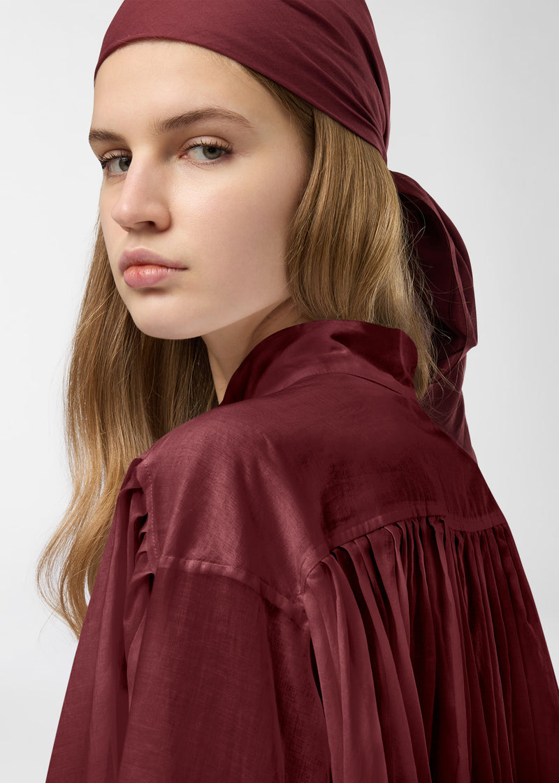 Detail view — Rania Shirt: Model looks over shoulder, showing mandarin collar, pleated back yoke and flowing semi-sheer rouge noir cotton muslin drape against a neutral studio background.